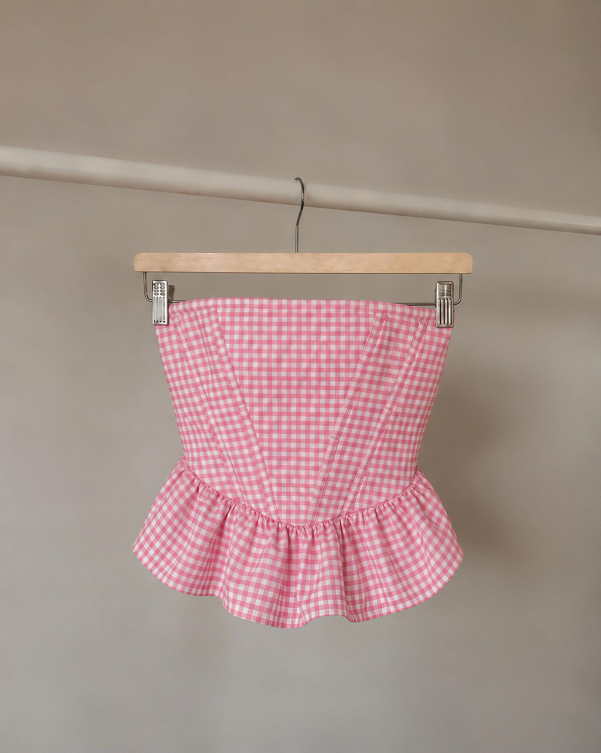 A pink and white gingham strapless corset-style top with a ruffled peplum hem, hanging from a wooden hanger against a plain beige wall.