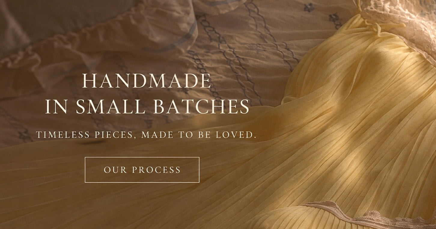 A close-up of a flowing pale yellow fabric with a ruffled, pleated texture, laid over a beige patterned textile. White text on the left reads 'HANDMADE IN SMALL BATCHES' with smaller tagline text below and a button reading 'OUR PROCESS'.